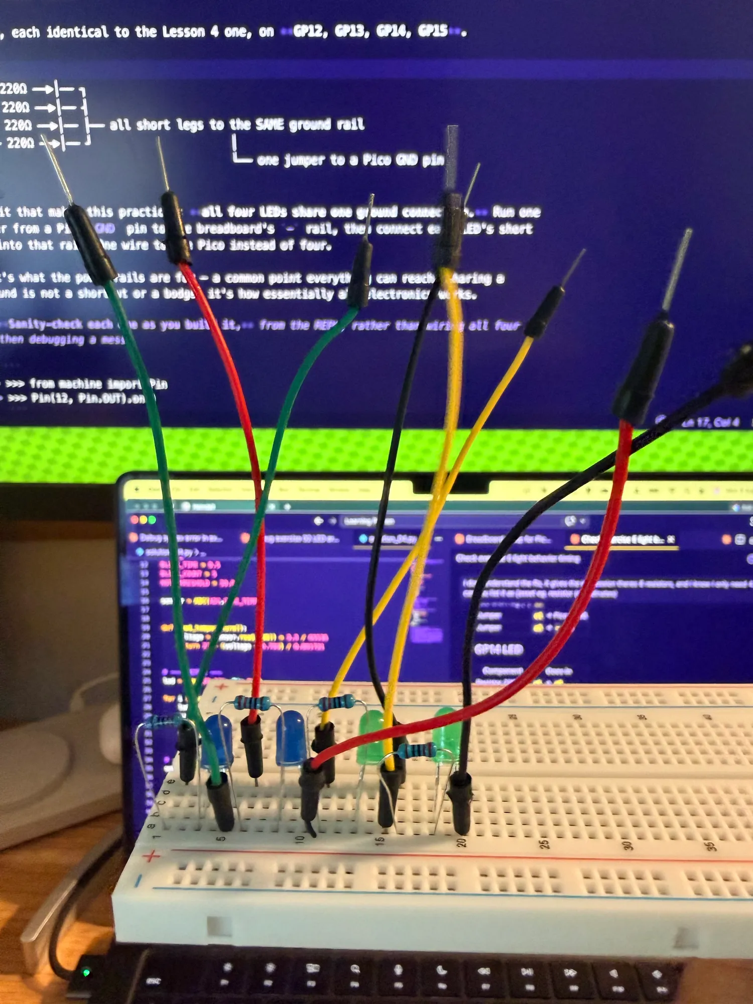 A bread board with wires sticking out of it, unplugged and 4 LEDs, one of which is sort of lit. Sitting in front of a purple screen of VS code, blurry in the bakground.